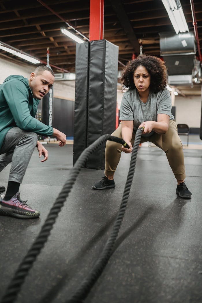 services-03 Strong African American female in activewear training with battle ropes near fit male coach in fitness center