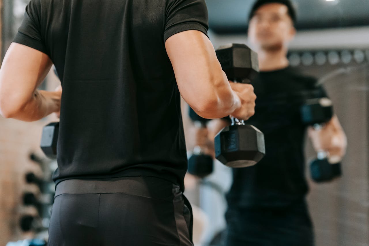 services-02 Back view of unrecognizable male in black activewear doing dumbbell curls while standing near mirror during workout on blurred background